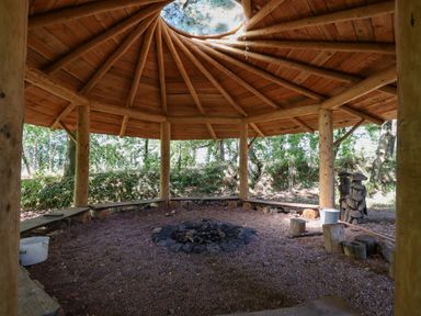 A wooden gazebo with a central fire pit and benches surrounded by trees at Nethercott House in Iddesleigh near Hatherleigh