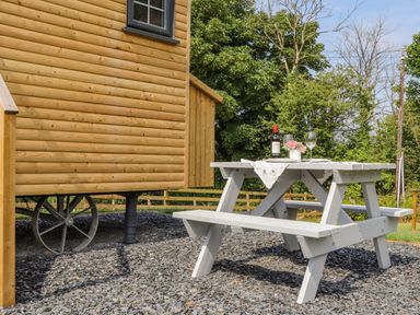 A wooden picnic table with a bottle of wine glasses and flowers next to a wooden wagon in a garden at Rose in Illogan