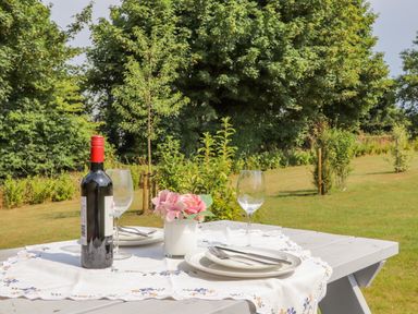 An outdoor picnic table set with plates forks wine glasses a bottle of wine and a flower vase in a garden