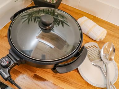 An electric skillet with a glass lid on a wooden countertop next to a rolled towel and utensils on a plate at Rose in Illogan