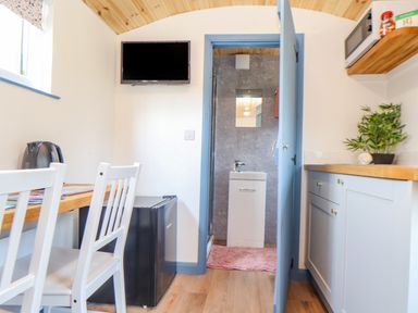 A small kitchen area with white chairs a wooden table a black kettle a mini fridge and a bathroom visible through a blue door at Rose in Illogan
