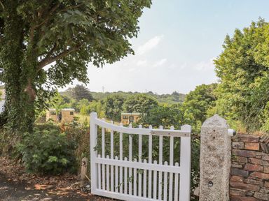 A white wooden gate opening to a path with trees and buildings in the distance at Rose in Illogan