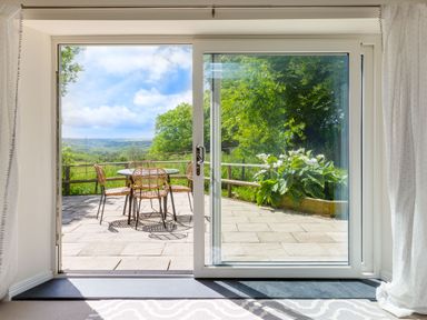 A patio with a round table and four chairs surrounded by plants and trees seen through sliding glass doors at Goose Cottage within the Helland Barton Farm collection in Delabole
