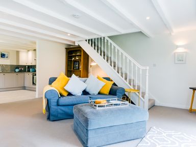 A living room with blue sofa and yellow cushions next to stairs and an open kitchen in the background at Goose Cottage within the Helland Barton Farm collection in Delabole