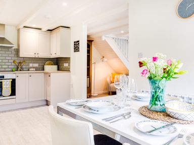 A kitchen and dining area with a set table and flowers in a vase with a small seating area under stairs at Goose Cottage within the Helland Barton Farm collection Delabole