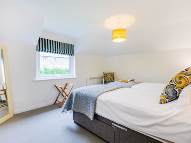A bedroom with twin beds blue quilted bed covers wooden luggage rack and a window with striped blind at Goose Cottage within the Helland Barton Farm collection Delabole