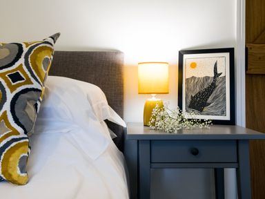 A bedside table with a lamp flowers and a framed artwork next to a bed with a patterned pillow at Goose Cottage within the Helland Barton Farm collection Delabole