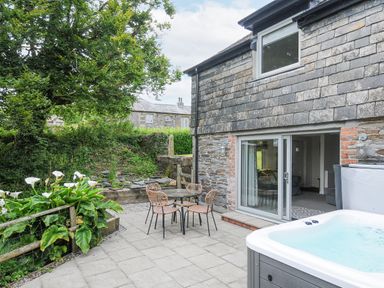 A patio with a table and chairs next to a stone building with sliding doors and a hot tub at Goose Cottage within the Helland Barton Farm collection Delabole