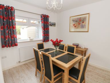 A dining room with a wooden table and six chairs a vase of red flowers on the table floral curtains a window a small wooden side table with candles and a bowl and a framed picture on the wall at Sandtop in Tenby