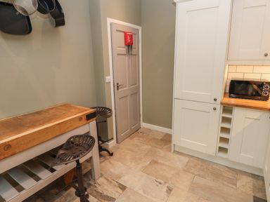 A kitchen with a wooden table and two stools a door with a fire alarm white cabinets and a microwave on the counter at Sandtop in Tenby