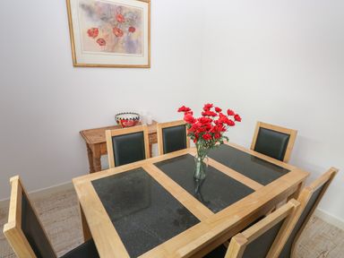 A dining room with a wooden table with black panels and six chairs a vase with red flowers on the table a wooden side table with a bowl and three small white containers and a framed floral painting on the wall at Sandtop in Tenby