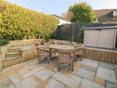 A backyard patio with a round wooden table and four wooden chairs surrounded by wooden raised beds with plants and a storage shed at Sandtop in Tenby