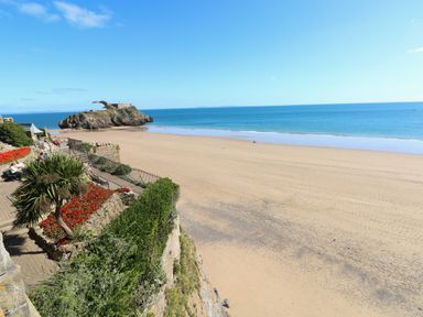 A sandy beach with a rocky island and a garden with flowers and benches overlooking the sea at Sandtop in Tenby