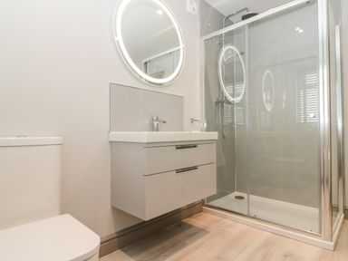 A bathroom with a toilet a wall-mounted sink with drawers a round illuminated mirror and a glass shower enclosure at 41 Chatsworth Gardens in Scarborough