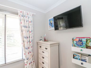 A corner of a room with a window with sea-themed curtains a white chest of drawers a wall-mounted TV and white shelves with books at 41 Chatsworth Gardens in Scarborough