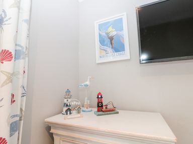 A corner with a TV mounted on the wall a framed north east coast poster a decorative bird a red lighthouse figurine a blue and white lighthouse figurine and a small beach hut decoration on a white cabinet with patterned curtains at 41 Chatsworth Gardens in Scarborough