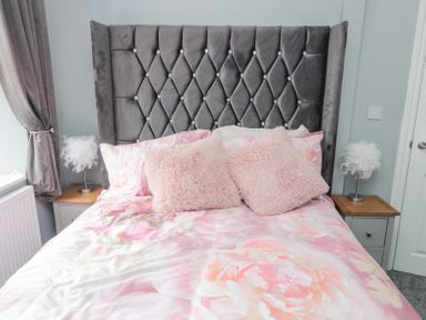A bedroom with a bed with a grey tufted headboard two pink pillows floral bedding and two bedside tables with feather lamps at 4 Tyne View in Wark