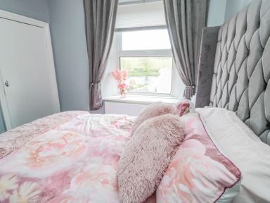 A bedroom with a grey tufted headboard a bed with floral bedding and pink pillows a window with grey curtains and a vase with flowers at 4 Tyne View in Wark