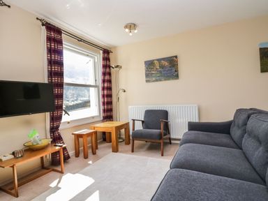 A living room with a gray sofa armchair wooden tables a TV and window with striped curtains at Fellside View in Chapel Stile