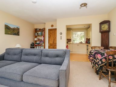 A living room with a gray sofa, wooden bookshelf, wooden dining table with patterned tablecloth, and a view into a kitchen at Fellside View in Chapel Stile