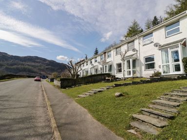 A row of white houses with glass porches along a sloped grassy area with stone steps and a road with parked cars at Fellside View in Chapel Stile