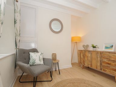 A corner of a room with a gray armchair and a cushion beside a small wooden side table a round wall mirror a wooden sideboard with a lamp plant and a framed picture on top at Carvannel Cottages North Cliffs near Portreath