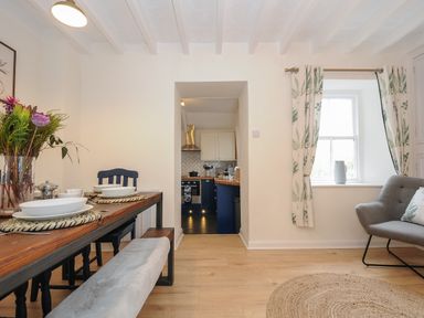 A dining table with plates and flowers beside a doorway leading to a kitchen in a room with a window and gray sofa at Carvannel Cottages North Cliffs near Portreath
