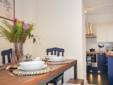 A dining table with plates and a vase of flowers next to a kitchen with blue cabinets and a stove at Carvannel Cottages North Cliffs near Portreath