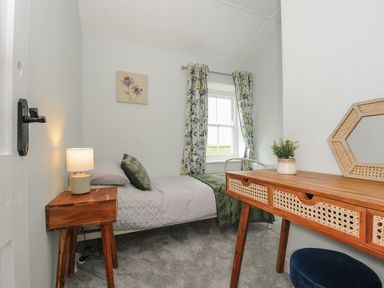 A small bedroom with a single bed a wooden bedside table with a lamp a wooden desk with drawers a plant and a mirror patterned curtains and a window at Carvannel Cottages North Cliffs near Portreath