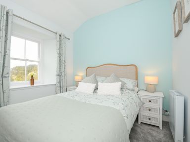 A bedroom with a double bed bedside tables lamps a window with curtains and flowers on the sill at Carvannel Cottages North Cliffs near Portreath