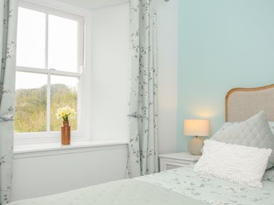 A bedroom with a bed a nightstand with a lamp a window with curtains and a vase with flowers at Carvannel Cottages North Cliffs near Portreath