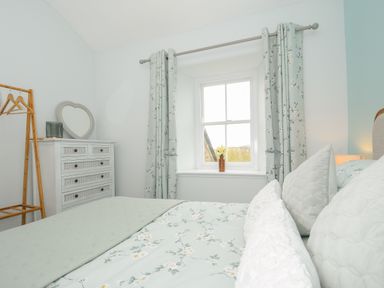 A bedroom with a bed with floral bedding a dresser a heart shaped mirror and a window with floral curtains at Carvannel Cottages in North Cliffs near Portreath