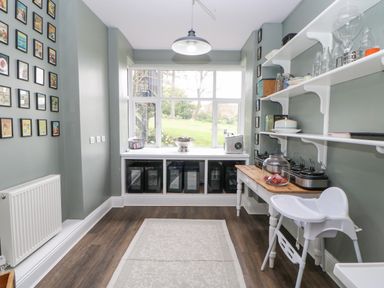 A pantry or utility room with shelves holding glassware and containers a wooden table with a glass jar and multiple slow cookers a high chair a wall with framed pictures and a window showing a garden outside at Heatherbrae in Middleton-In-Teesdale