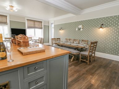 A dining area with a wooden table set with plates and glasses wooden chairs and a bench and a kitchen island with cutting boards and a bottle of prosecco at Heatherbrae in Middleton-In-Teesdale