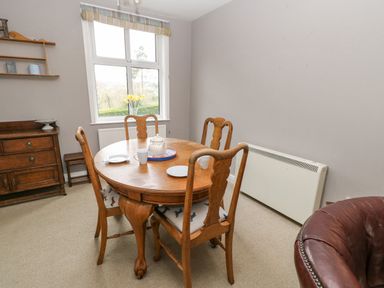 A dining room with a round wooden table and four chairs near a window with flowers on the sill at Heatherbrae in Middleton-In-Teesdale