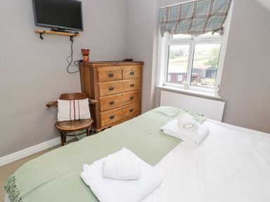 A bedroom with a bed covered in white and green linens a wooden chair with a cushion a wooden dresser a window with a plaid curtain and a wall-mounted TV at Heatherbrae in Middleton-In-Teesdale