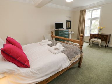 A bedroom with a wooden bed with red pillows and folded towels a green chest of drawers with a television a basin a wooden desk with a stool and flowers by a window at Heatherbrae in Middleton-In-Teesdale