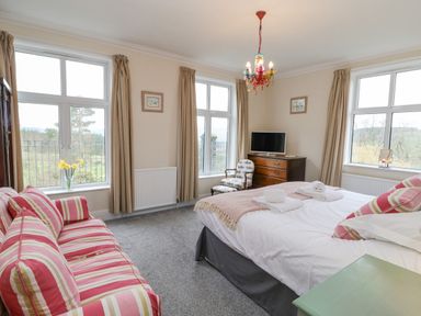 A bedroom with a striped sofa a bed with folded towels a wooden dresser with a television and windows with curtains at Heatherbrae in Middleton-In-Teesdale