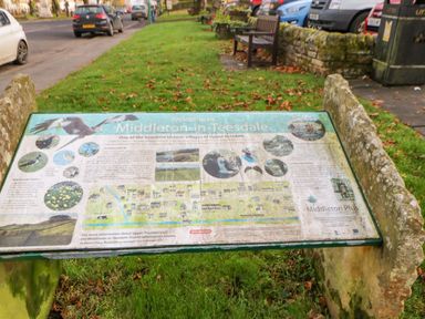 An outdoor information board about Middleton-in-Teesdale on a grassy area near a road with parked cars and a bench