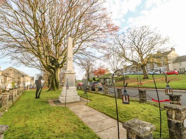 A memorial obelisk with metal soldier silhouettes and lanterns on a grassy area near a road and houses at Heatherbrae in Middleton In Teesdale