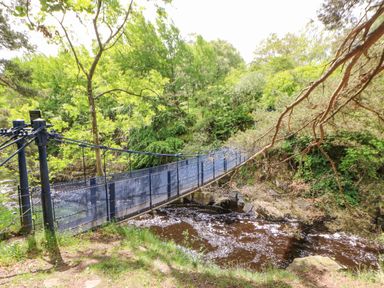 A metal suspension bridge over a river surrounded by green trees at Heatherbrae in Middleton-In-Teesdale