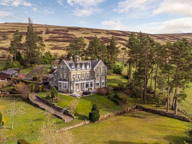 A large stone house surrounded by trees and grass with hills in the background at Heatherbrae in Middleton-In-Teesdale