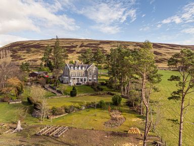 A large stone house surrounded by trees and garden beds with a hill in the background at Heatherbrae in Middleton-In-Teesdale