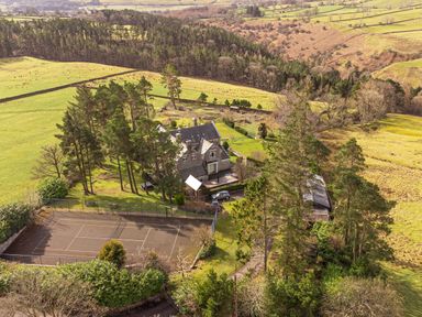 An aerial view of a house surrounded by trees and fields with a tennis court in the foreground at Heatherbrae in Middleton In Teesdale