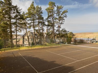 An outdoor tennis court with a net surrounded by trees and a stone building in the background at Heatherbrae in Middleton-In-Teesdale