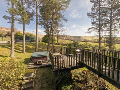 An outdoor wooden deck with a table and chairs next to a hot tub in a garden with trees and hills in the background at Heatherbrae in Middleton-In-Teesdale