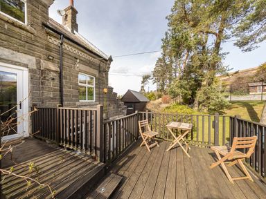 A wooden deck with two chairs and a table outside a stone building with trees and grass in the background at Heatherbrae in Middleton-In-Teesdale