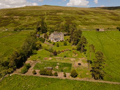 A large stone house surrounded by trees and garden plots on a hillside at Heatherbrae in Middleton-In-Teesdale