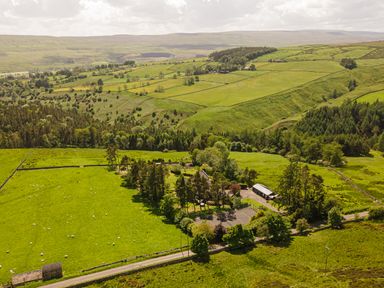 Green fields with sheep and a cluster of buildings surrounded by trees in a rural landscape at Heatherbrae in Middleton-In-Teesdale