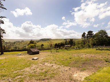 An outdoor fenced garden area with small trees and wooden structures overlooking rolling hills and cloudy sky at Heatherbrae in Middleton-In-Teesdale